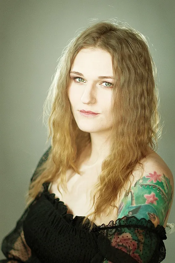 Kobieta o jasnych falowanych włosach, w czarnej koronkowej bluzce, z tatuażem na ramieniu