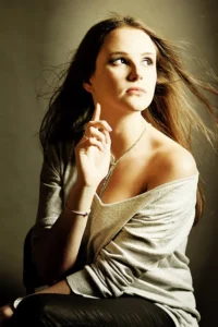 Kobieta w szarej bluzce, delikatnie dotykająca włosów, patrzy w bok z zamyślonym wyrazem twarzy