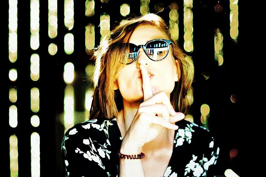 Kobieta w okularach przeciwsłonecznych, przykładająca palec do ust w geście ciszy