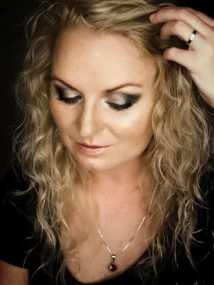 Portret kobiety o blond kręconych włosach, z delikatnym makijażem i złotym naszyjnikiem