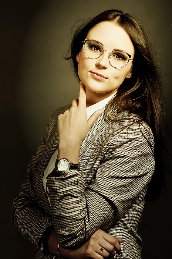 Młoda kobieta w okularach i stylowej marynarce, z delikatnym uśmiechem i subtelnym gestem dłoni