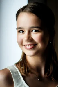 Młoda dziewczyna z uśmiechem, stojąca przy oknie, w naturalnym świetle