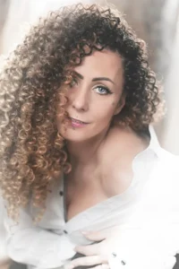 Kobieta z burzą kręconych włosów, w białej koszuli, patrząca uwodzicielsko