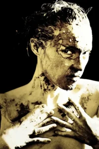 Kobieta pokryta gliną, patrząca intensywnie, w klimacie artystycznym i surowym